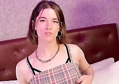 A bubbly trans woman with short brown hair playfully positions on a pink and white daybed, widening her legs, cupping her chest, and showing off her b